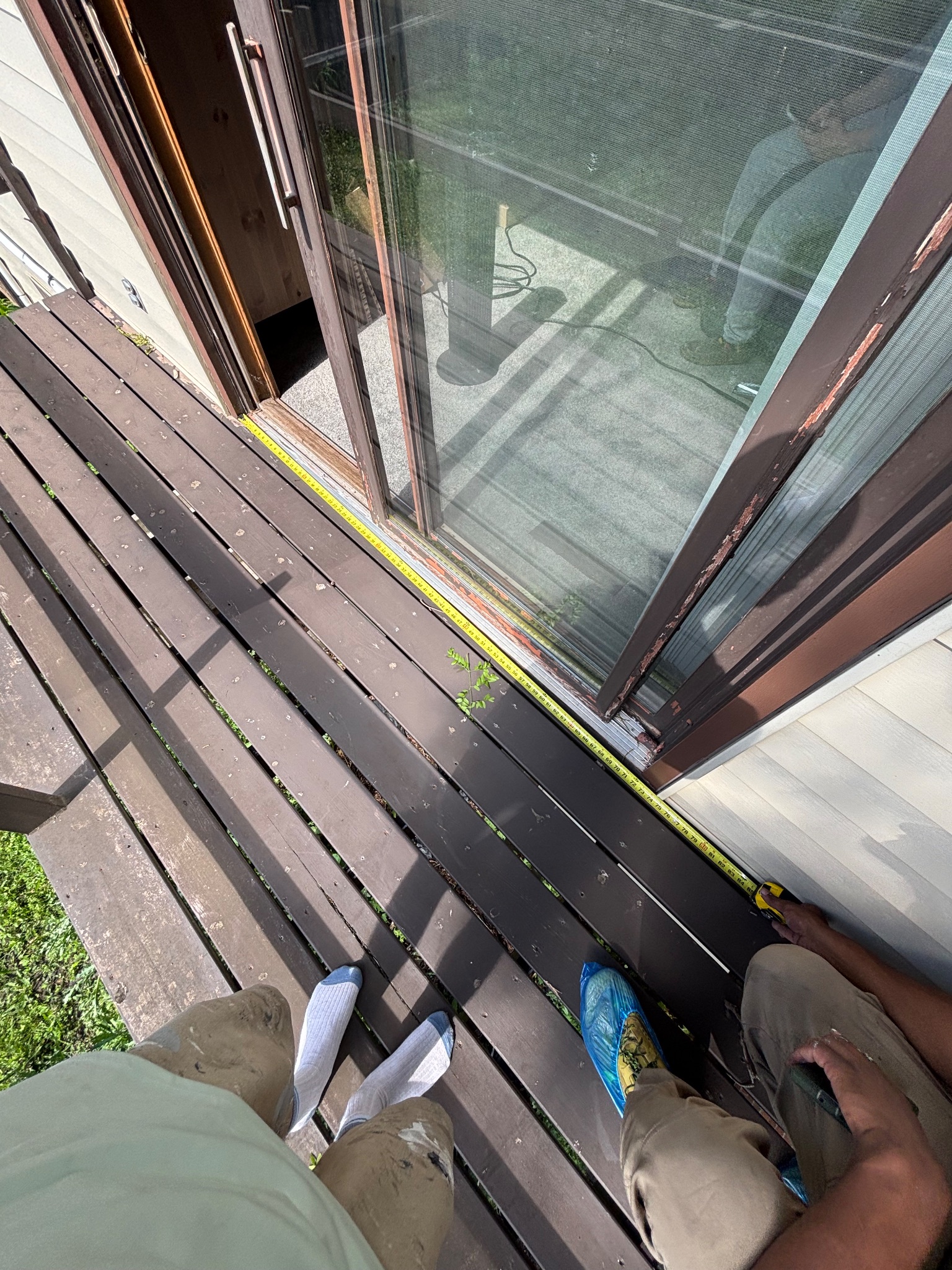 Measuring the exterior frame of an old sliding patio door in Ames, with a person's feet visible on a wooden deck, indicating preparation for replacement.