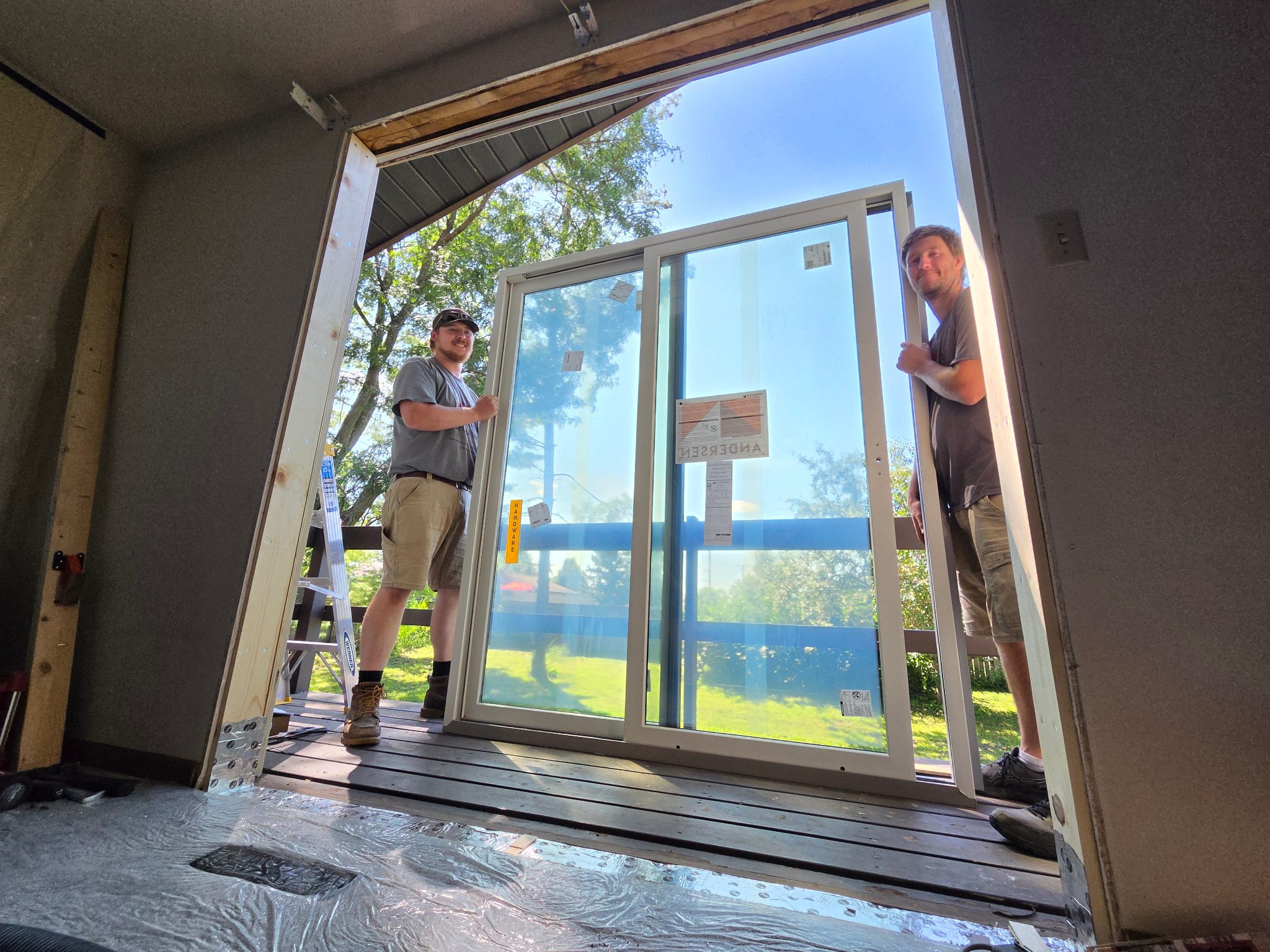 Two Iowa Home Consulting team members smiling while holding the new vinyl sliding patio door in Ames, ready for final placement into the prepared opening.