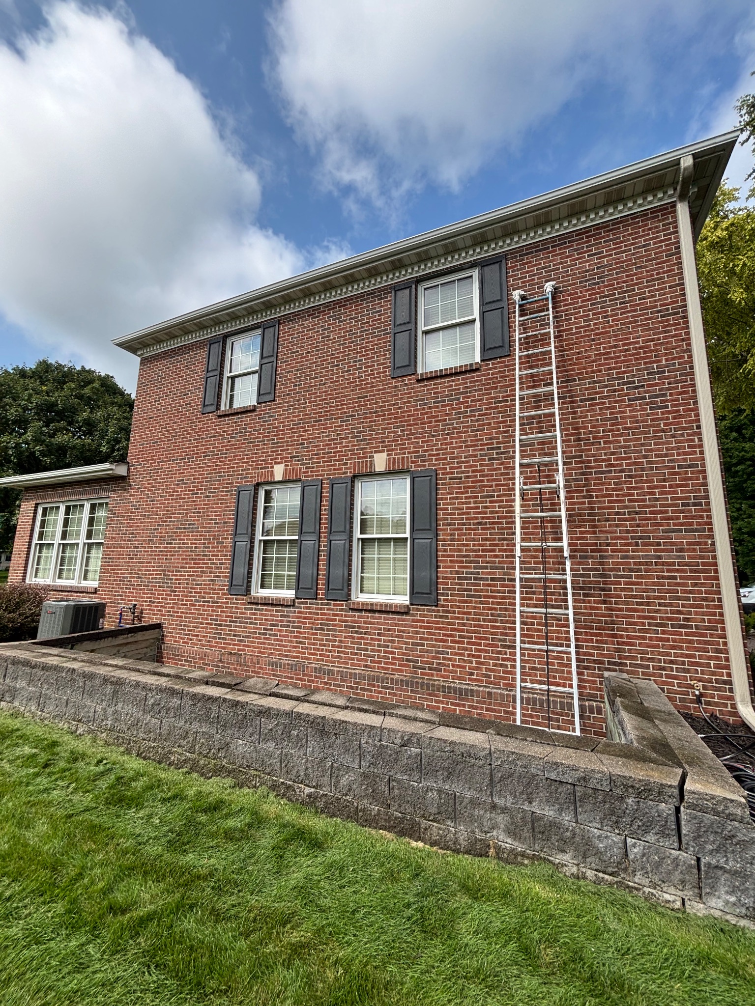 Side exterior of a red brick two-story home with reinstalled black shutters and a ladder leaning against the wall, Ames shutter reinstallation.