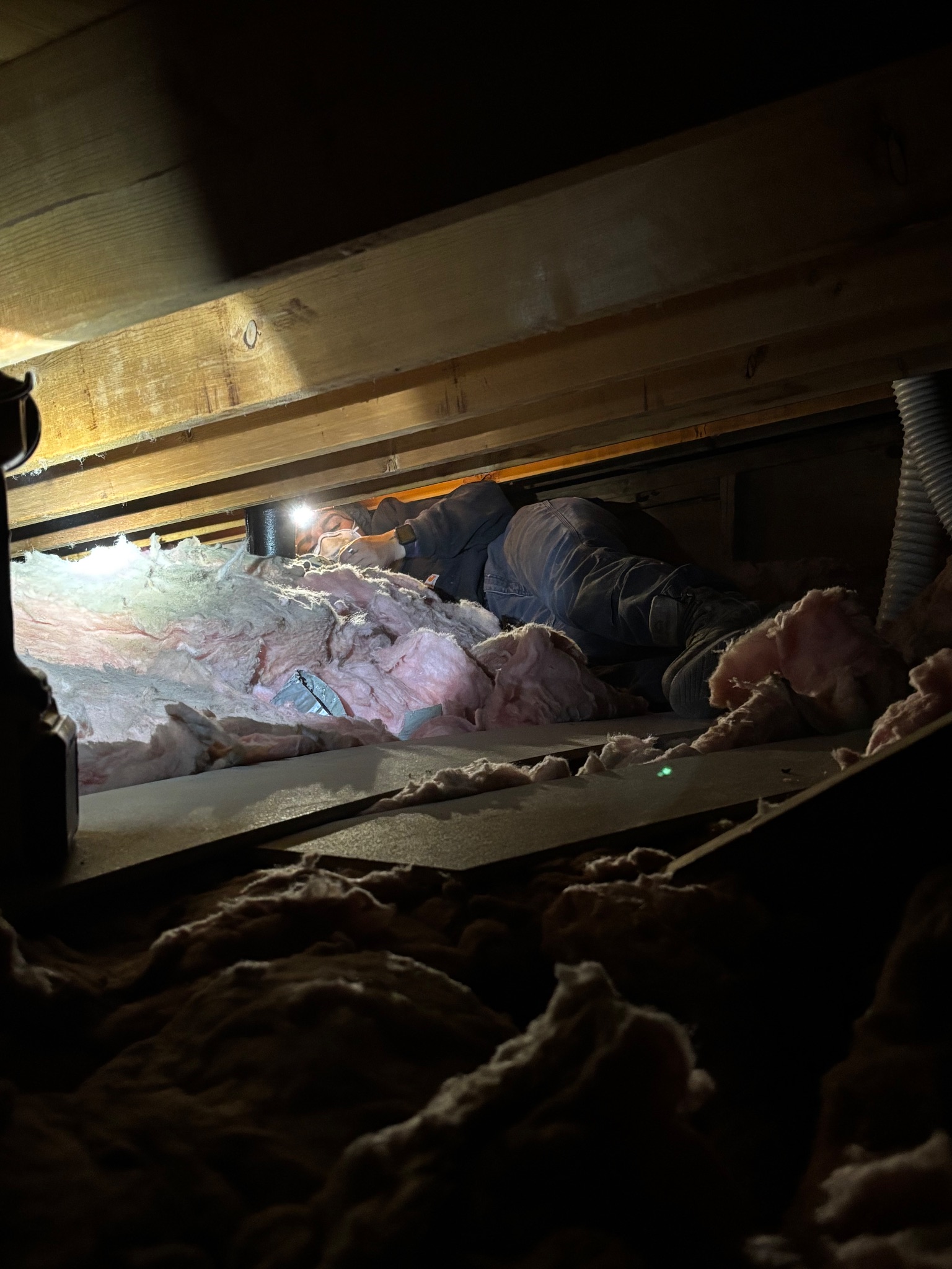 Handywoman working in a dimly lit attic, installing batt insulation around a bath fan vent line in Ames, Iowa.