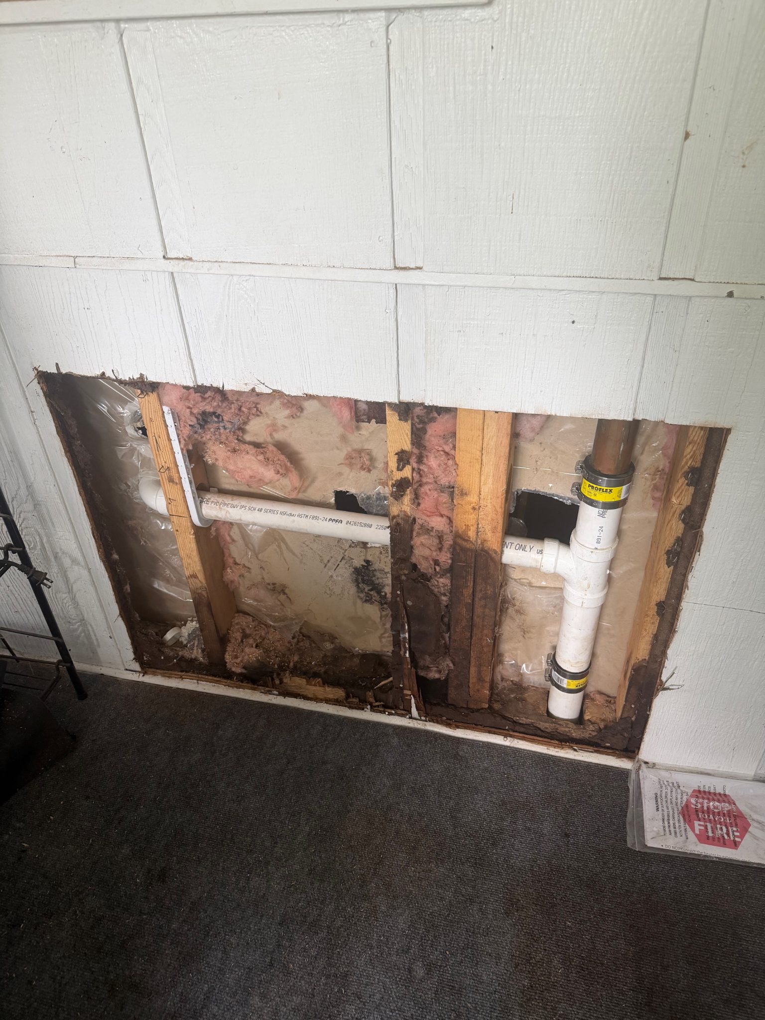 Damaged paneled wall exposing insulation and pipes in Ames, handyman repair.